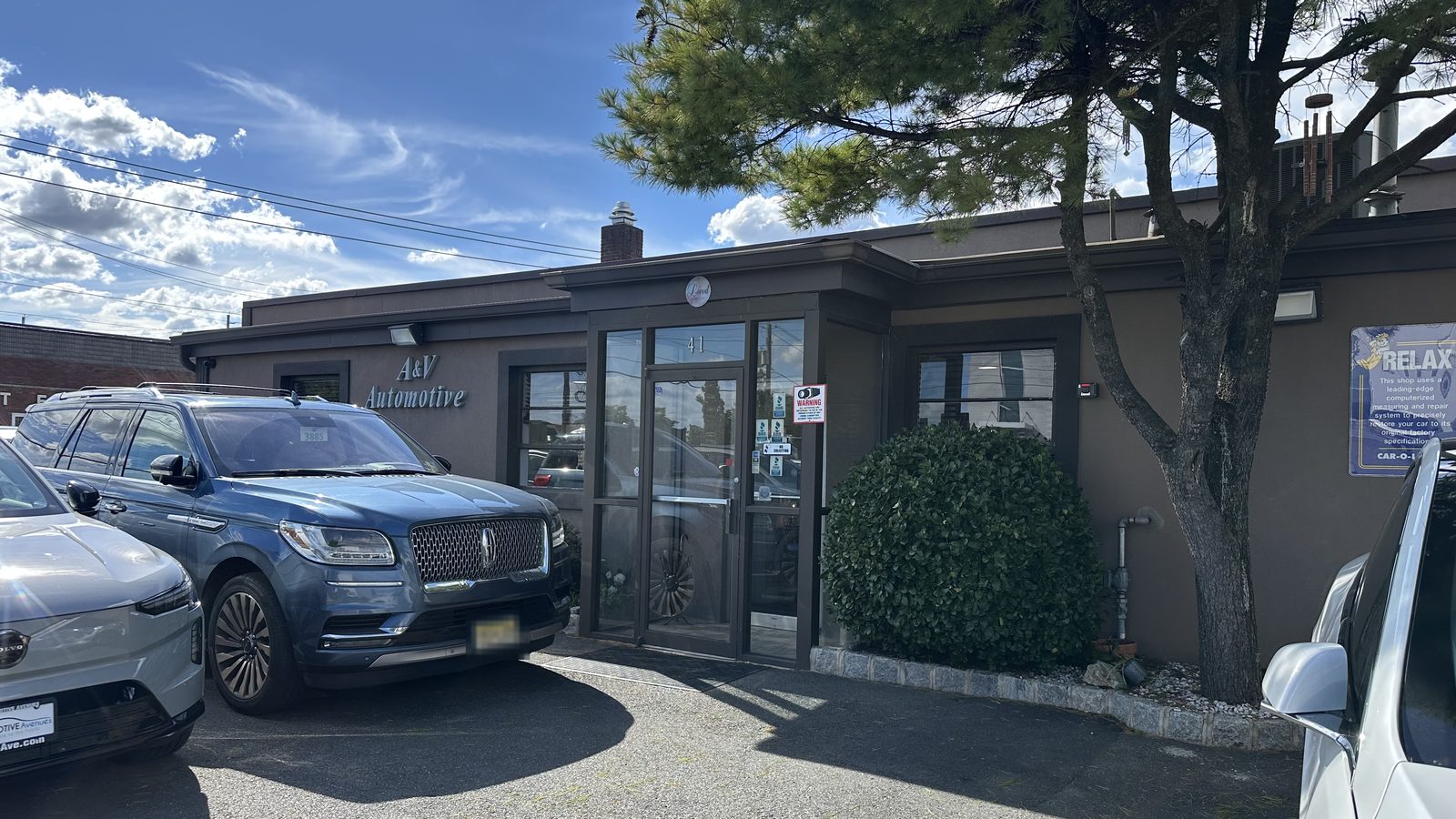 The front of A&V Automotive at 41 Romeo Street: a low brown building with the A&V Automotive sign beside the glass entrance, customer cars parked along the front under a pine tree