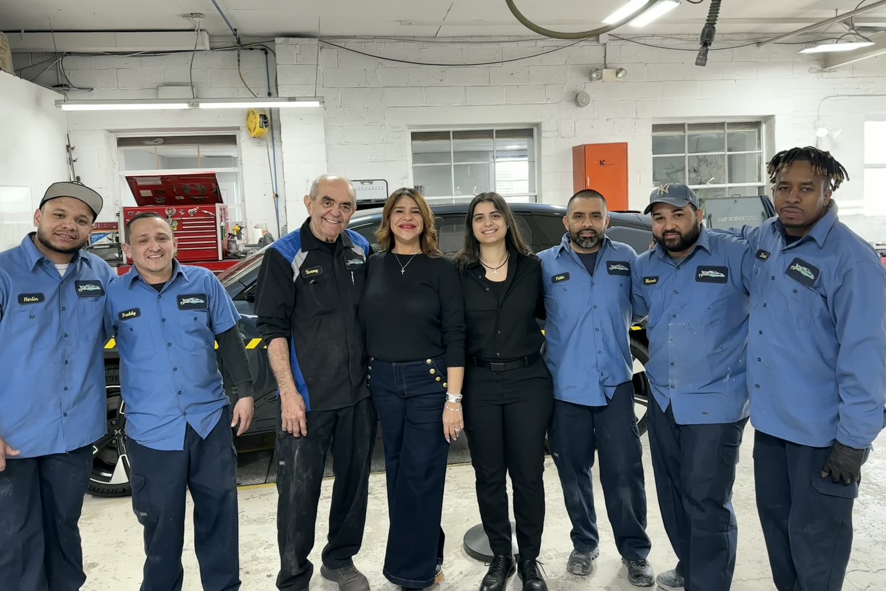 Venecia and the A&V Automotive crew, eight people standing arm in arm in the shop in front of a car on the lift